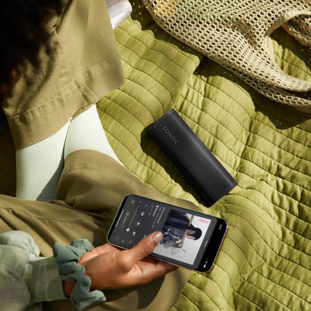 Sonos Roam 2 in Black outdoors on a picnic blanket with a person using the Sonos App
